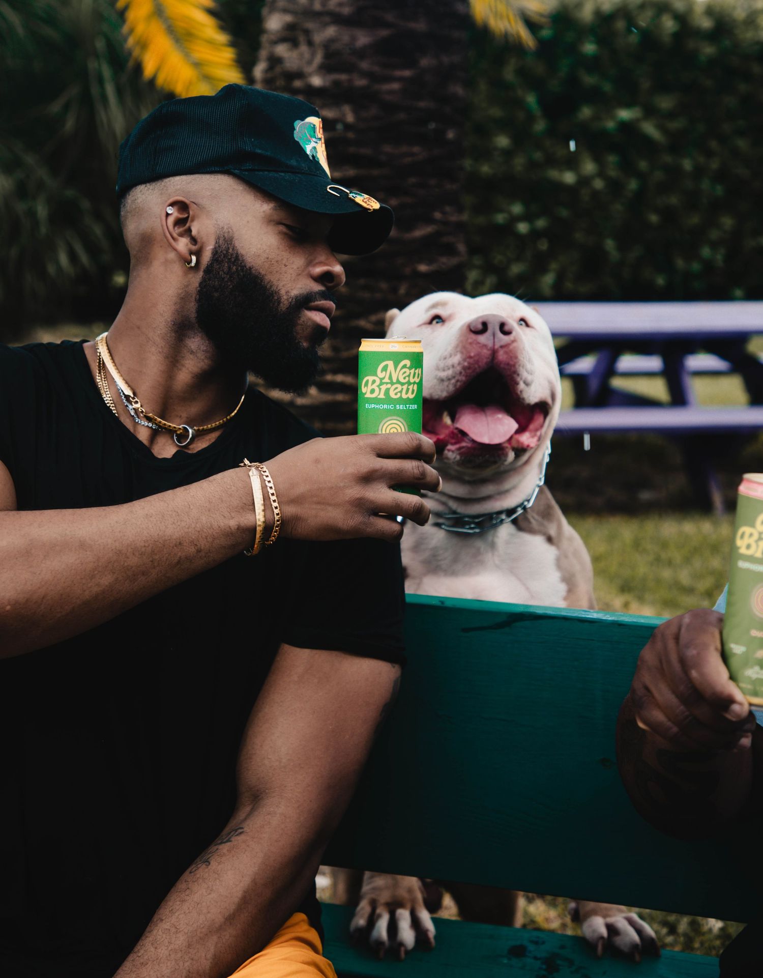 Person in baseball cap enjoys a drink outdoors with a happy white pit bull dog sitting beside them.