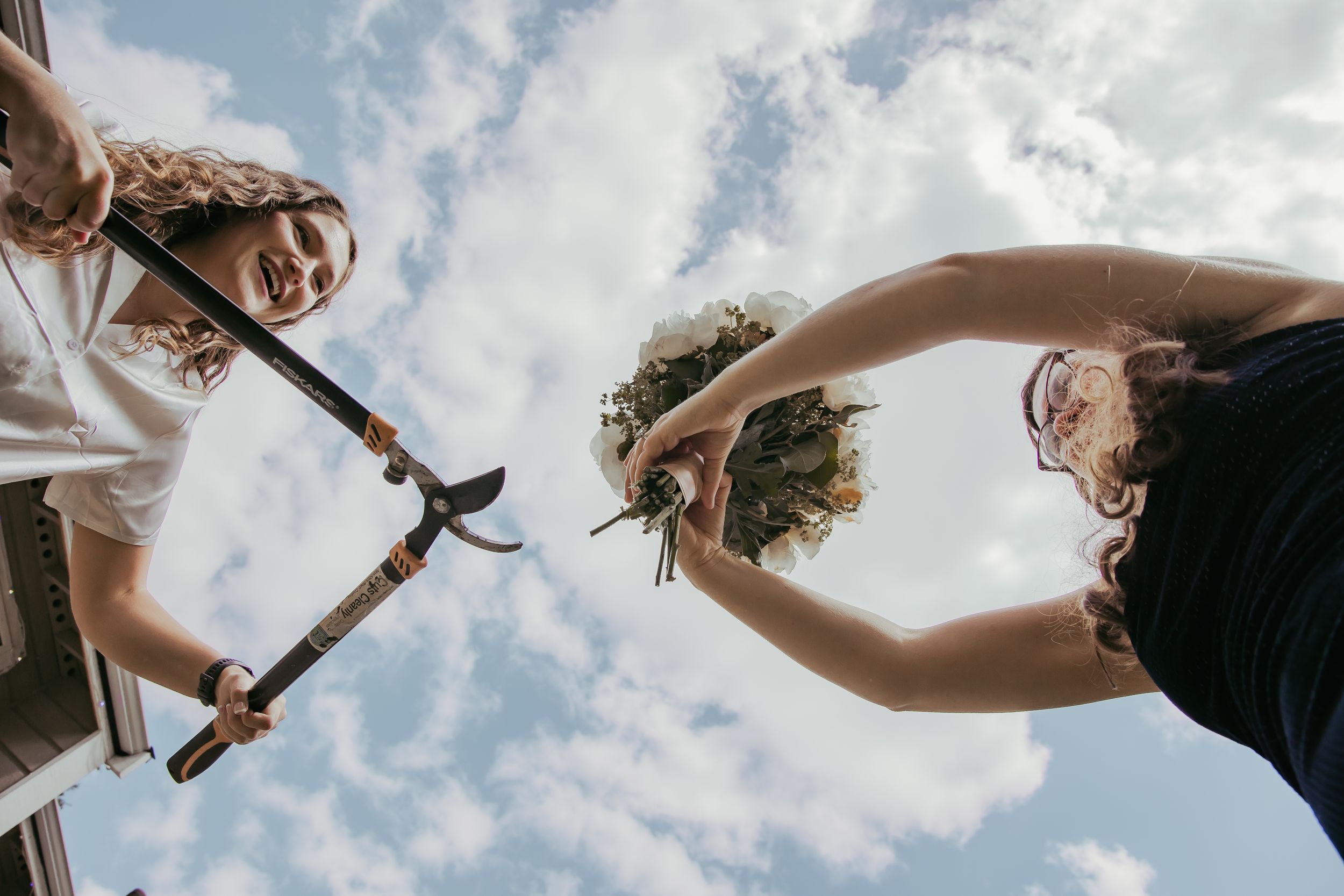 Low-angle view of two women outdoors, one holding garden shears and another holding a floral bouquet against a blue sky.