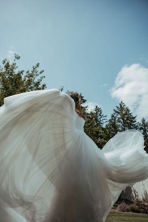 Bride in flowing white wedding dress billowing in the wind against a blue sky with trees in the background.