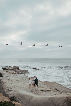 Family of three standing on rocky shore watching ocean waves with birds flying overhead on a cloudy day.