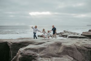 Family of four joyfully playing on rocky coastal cliffs with ocean waves in the background on a cloudy day.