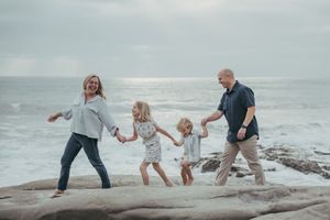 Family of four holding hands and walking on rocky beach with ocean waves in the background on a cloudy day.