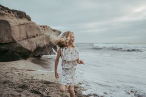 A young girl in a floral dress runs joyfully along a rocky beach with ocean waves in the background.