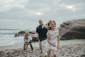 Family laughing and running on a sandy beach near rocky cliffs with ocean waves in the background.