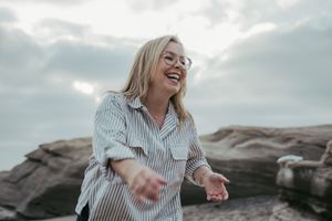 A joyful blonde woman in glasses and a striped shirt laughs outdoors near rocky cliffs under a cloudy sky.
