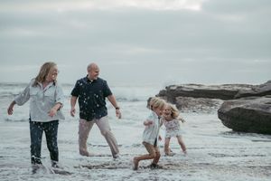 Family of four joyfully splashing in ocean waves on a rocky beach during an overcast day.