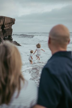 Two children playing in ocean waves at the beach while parents watch from behind on a cloudy day.
