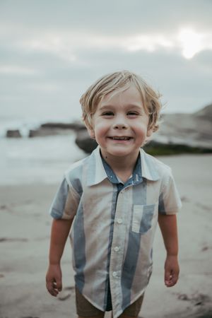 A smiling young blond boy in a plaid shirt stands on a sandy beach with rocky shores and cloudy sky.