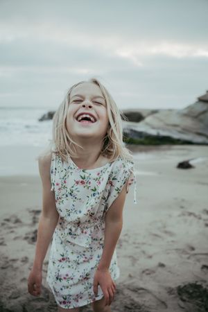 Joyful blonde girl in floral dress laughing on a sandy beach with ocean waves and rocks in background.