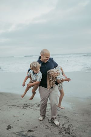 A bald father playfully carries two young children on a sandy beach with ocean waves in the background.