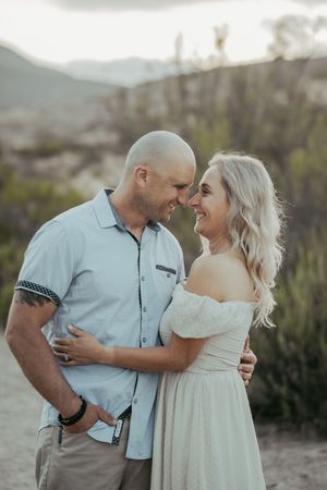A smiling couple embraces outdoors in a desert landscape, the bald man in a blue shirt and the blonde woman in a white off-shoulder dress.