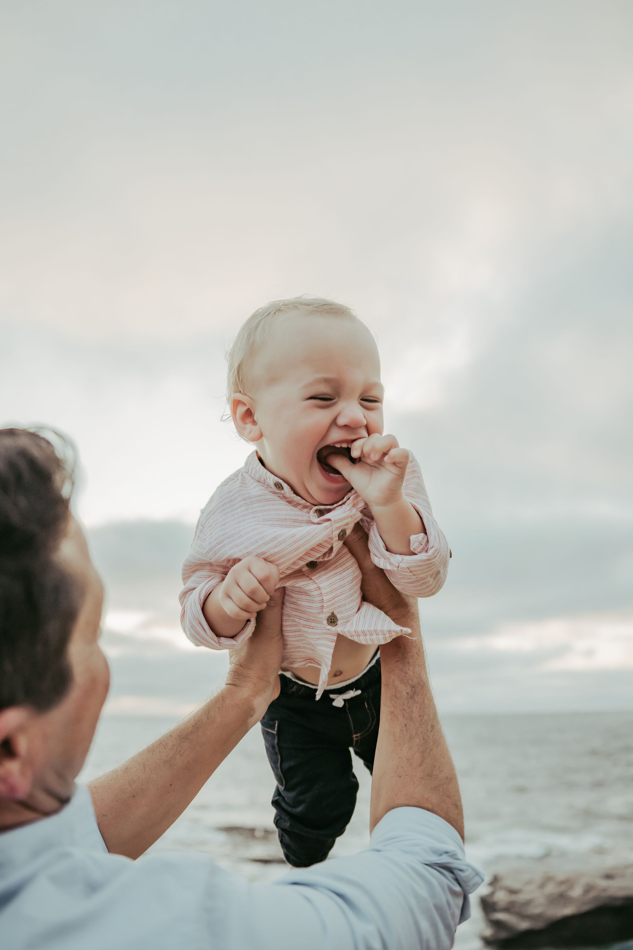 Father lifting laughing baby boy at the beach, cloudy sky in background.