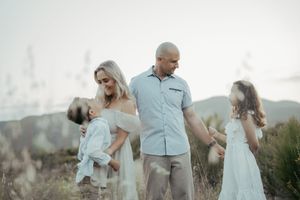 A family of four walks through a scenic field, parents holding children's hands in coordinated white and beige outfits.