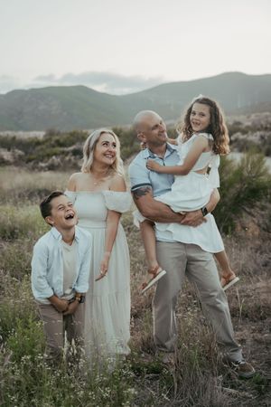 A happy family of four poses outdoors in a field with mountain views, dressed in white and neutral tones.