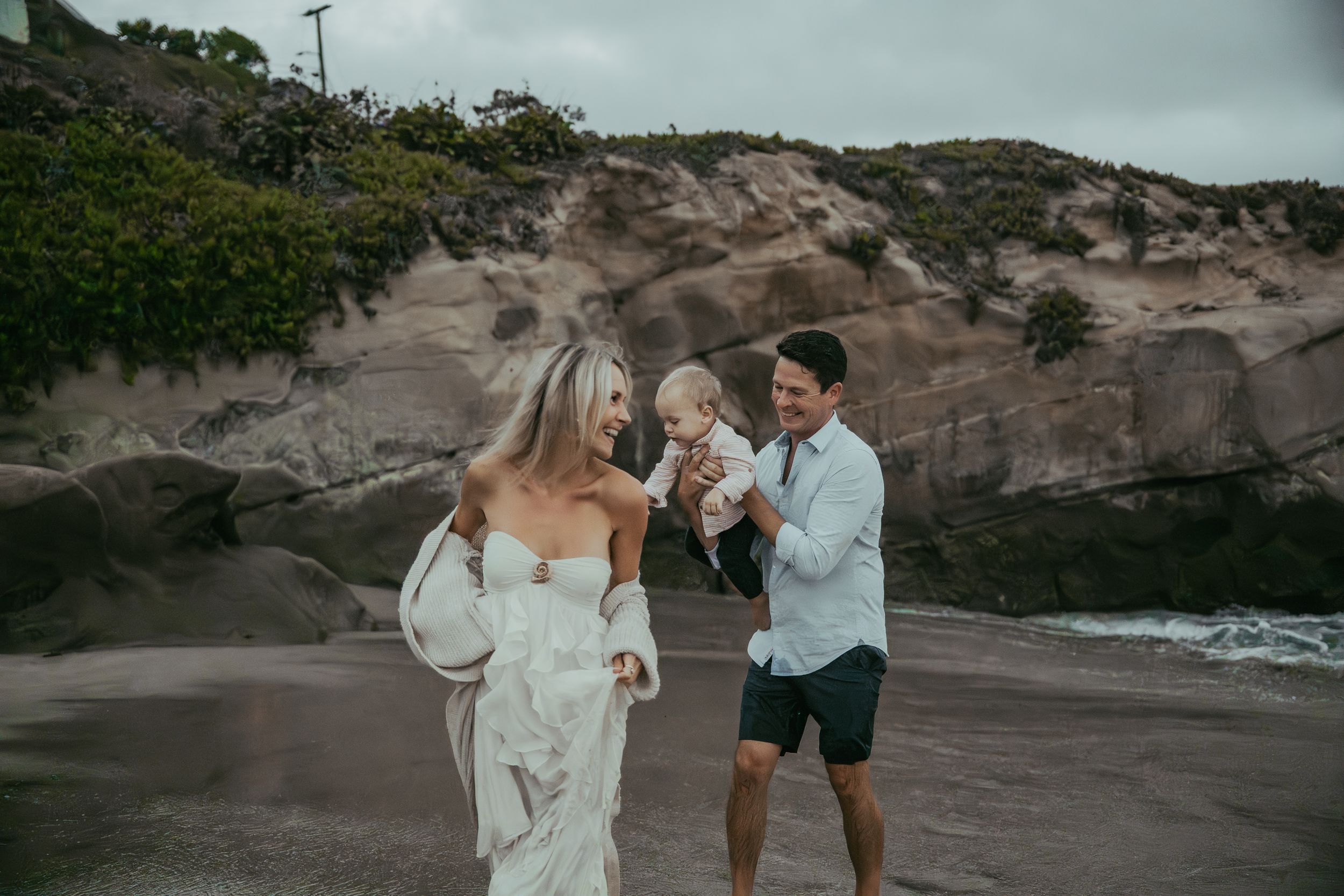 A couple in wedding attire holds their baby on a scenic beach with rocky cliffs in the background.