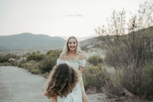 Two women in white dresses walk a desert path with mountains in the background at sunset.