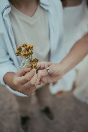 Two people holding small yellow wildflowers together, one wearing a silver ring, dressed in white and blue.