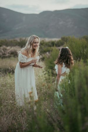 Two women in white dresses stand in a field with mountains, sharing a joyful moment surrounded by greenery.