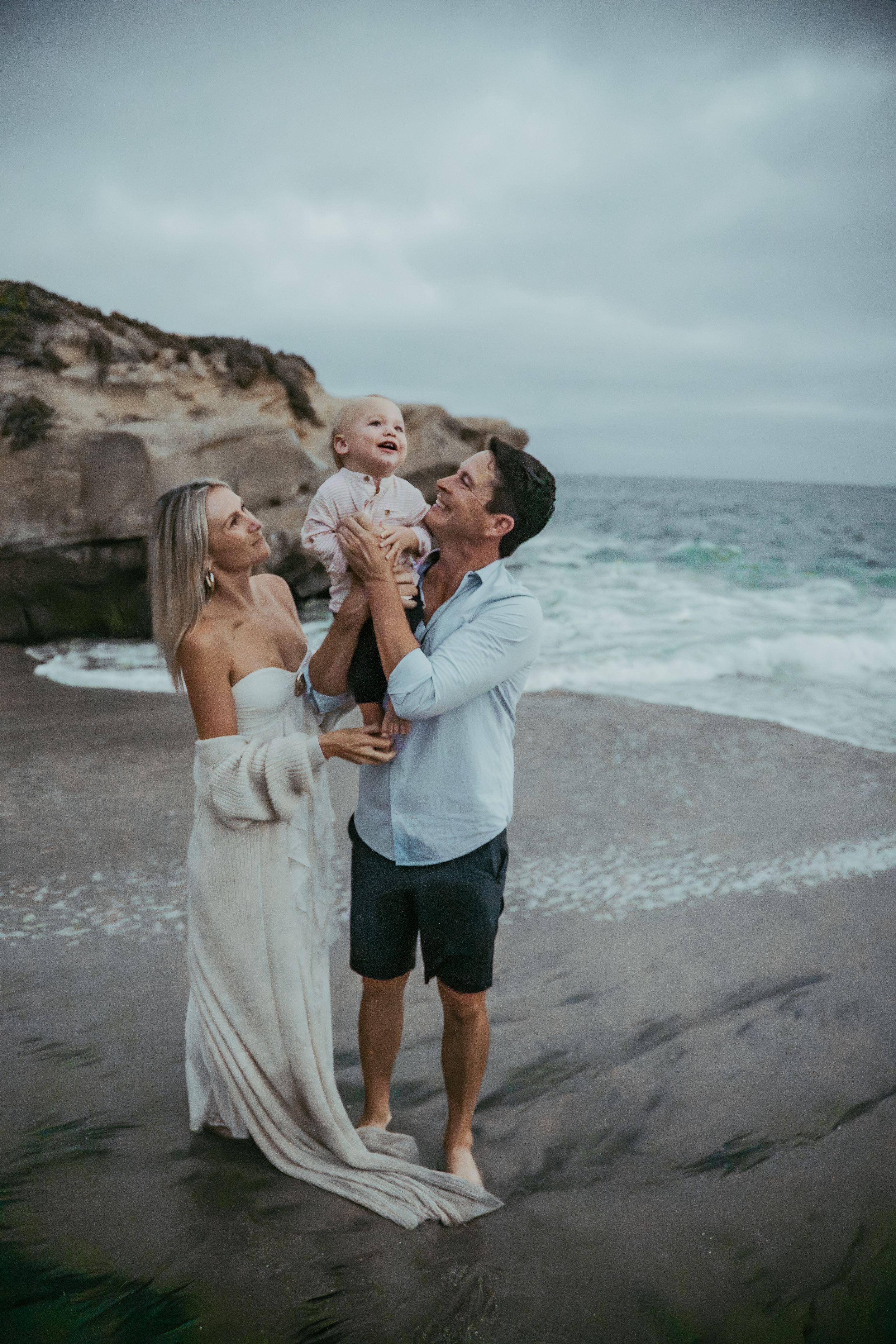 A joyful family of three poses on a rocky beach, with parents lifting their laughing baby against an overcast sky.