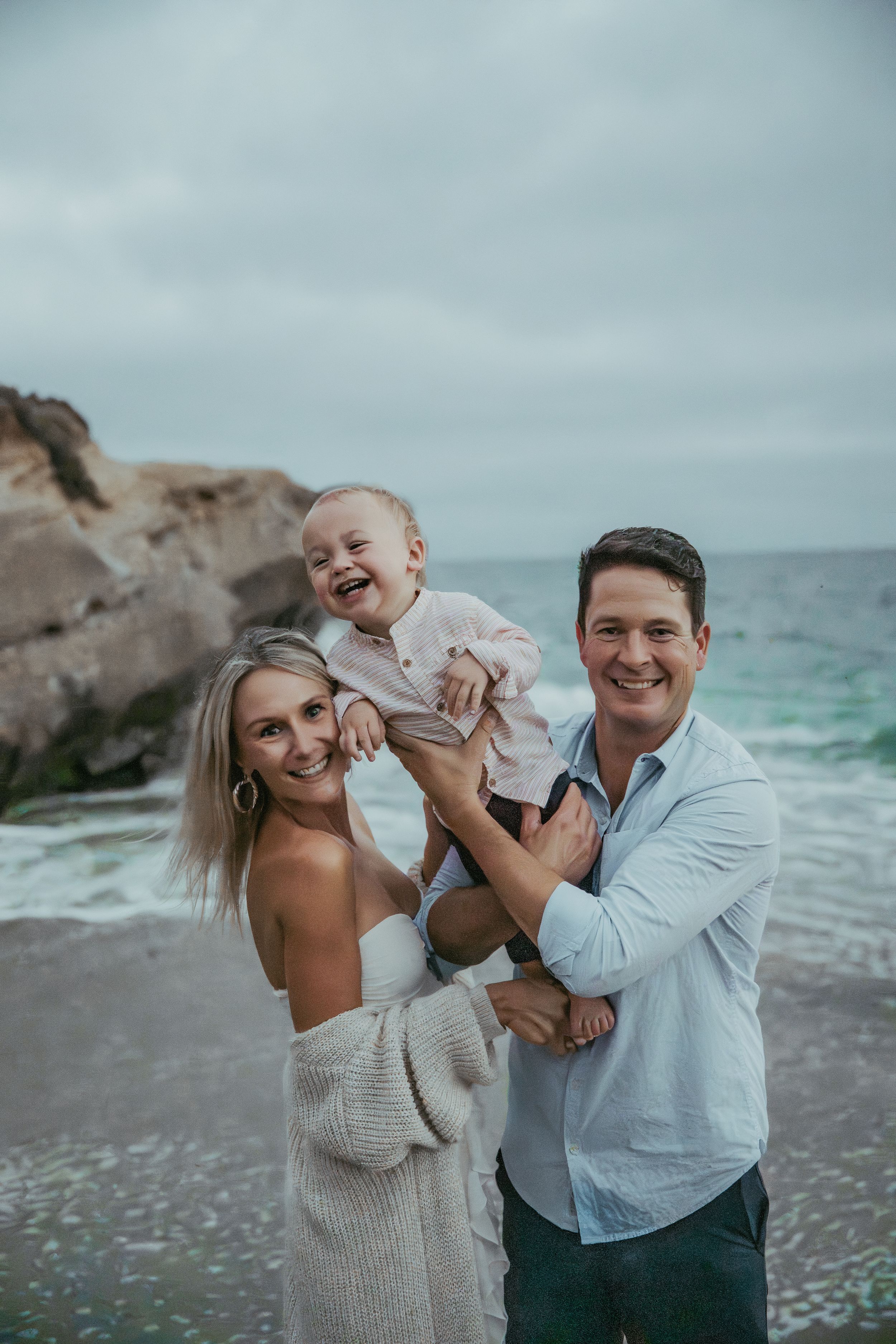 A happy family of three poses on a rocky beach, with parents holding their laughing toddler against an overcast sky.