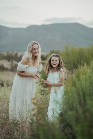 Two females in white dresses stand in a field with mountain backdrop, smiling joyfully together.