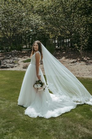 Bride in a flowing white ball gown with a long veil holding a bouquet, posing outdoors on green grass.