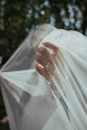 Bride's hand with diamond engagement ring elegantly draped in flowing white wedding veil outdoors.