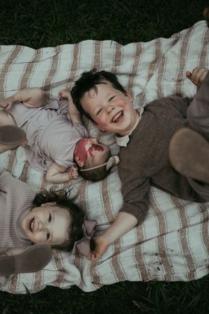 Two happy children laughing and lying on a striped picnic blanket on green grass outdoors.