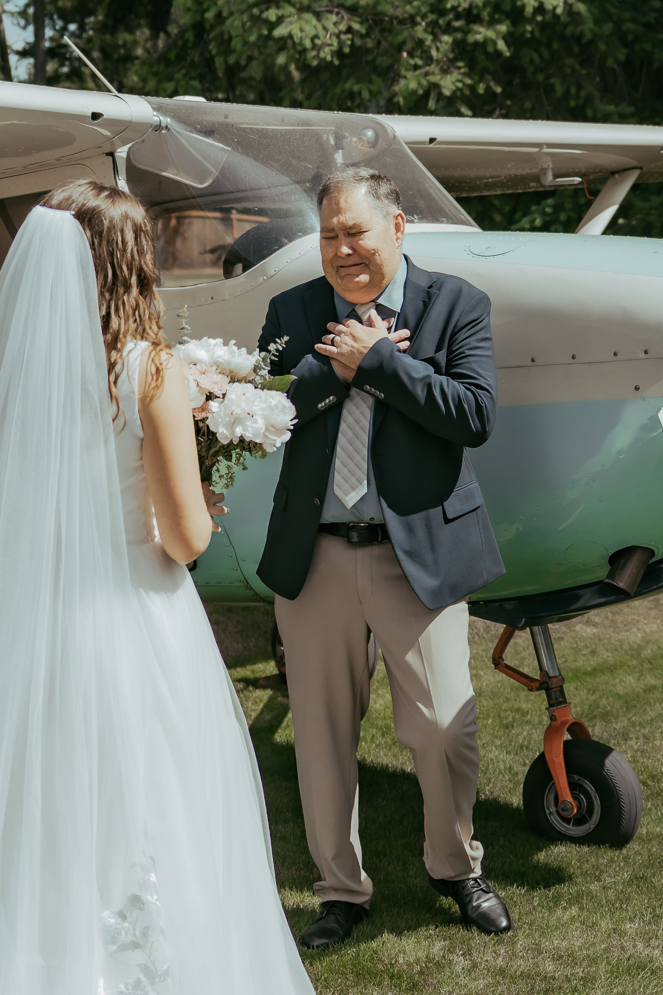 Emotional father reacts with joy seeing his daughter in wedding dress near a small plane outdoors.
