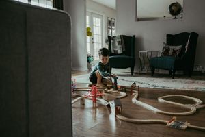 Young boy playing with wooden train tracks on living room floor surrounded by toy railroad set.