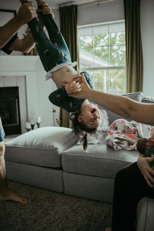 Parent playfully holding child upside down over couch while family laughs together indoors.