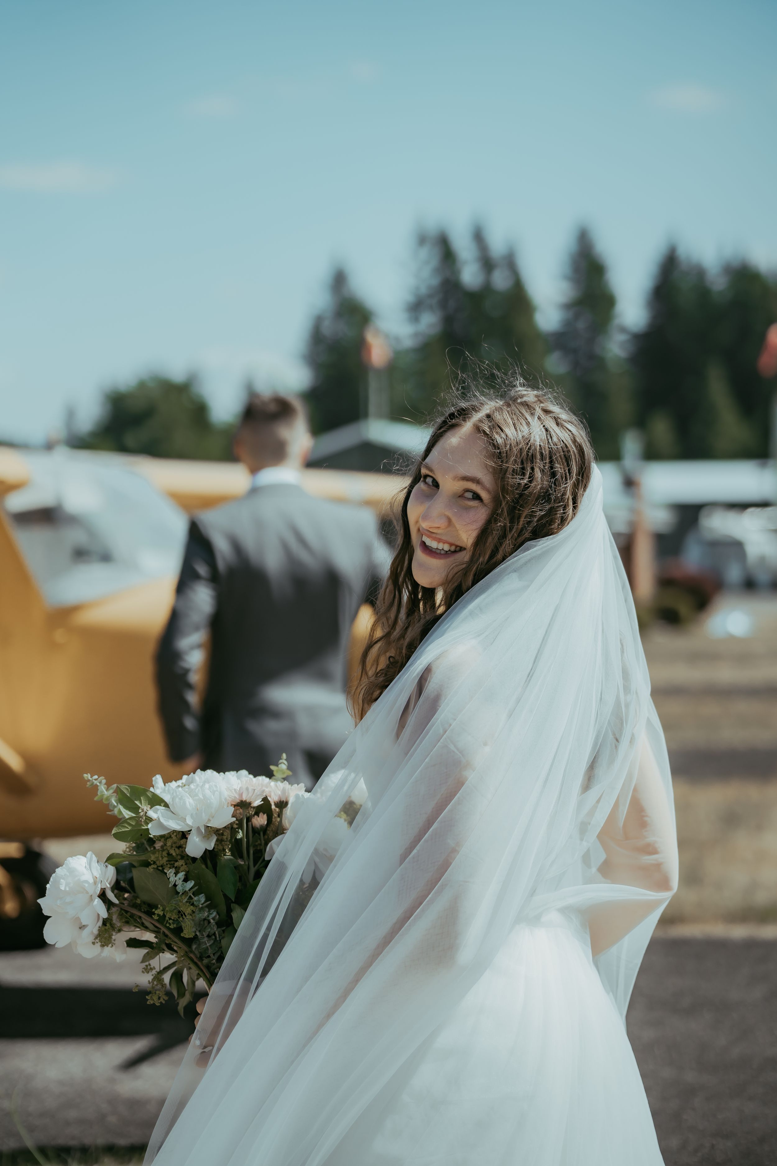 Smiling bride in white gown and veil holds bouquet at outdoor wedding venue with yellow plane in background.