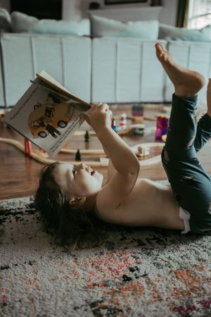 Young child lying on a rug reading a book with toys scattered on hardwood floor in background.