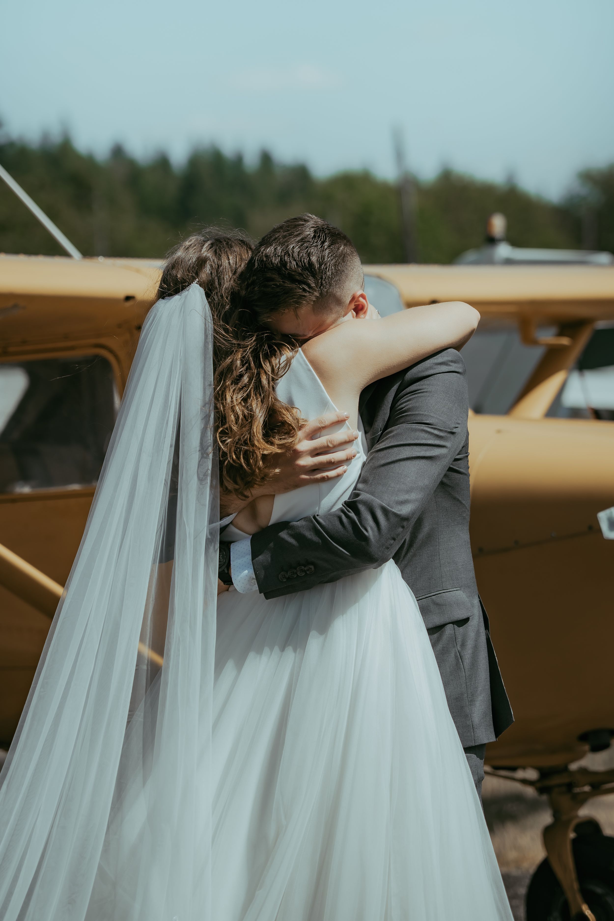 Bride and groom share romantic embrace near vintage yellow plane, bride wearing white gown and veil.