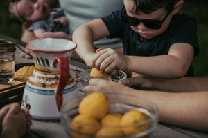 Boy in sunglasses squeezing a lemon at an outdoor table with a decorative pitcher and bowl of lemons.