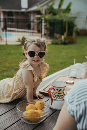 Smiling toddler girl in yellow dress wearing heart-shaped sunglasses at an outdoor lemonade stand.