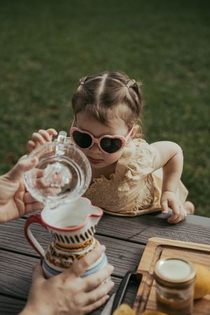 Toddler girl in heart sunglasses pouring water from glass pitcher at outdoor wooden table.