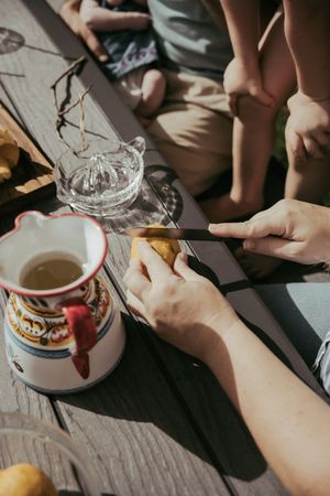 Hands cutting fruit with a knife on a wooden table with a decorative mug and glass juicer outdoors.