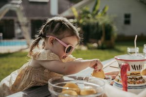Young girl in pink sunglasses reaches for food at an outdoor table near a pool on a sunny day.