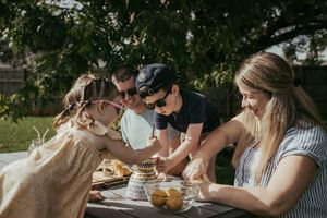 Happy family with two kids cutting cake together at outdoor picnic table under tree.