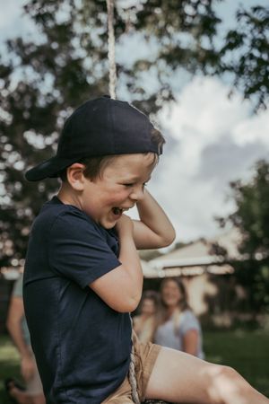 Happy young boy in a black cap laughing while sitting on a rope swing outdoors in a backyard.