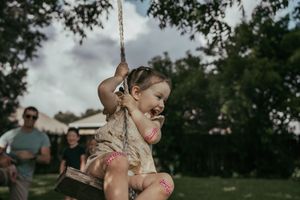 Joyful toddler girl swinging on a rope swing outdoors surrounded by trees and people.