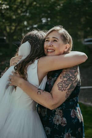 A bride in white hugs a smiling woman with floral tattoos in a dark floral dress outdoors.