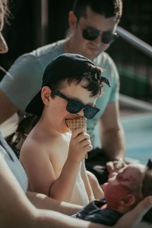Young boy wearing sunglasses and a cap eating an ice cream cone poolside with family nearby.