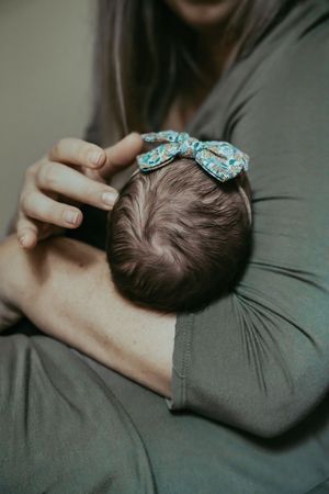 Mother cradling newborn baby with a floral bow headband against her chest in soft indoor light.
