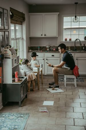 Father sits on chair using phone while two young children eat at small kitchen table in family home.