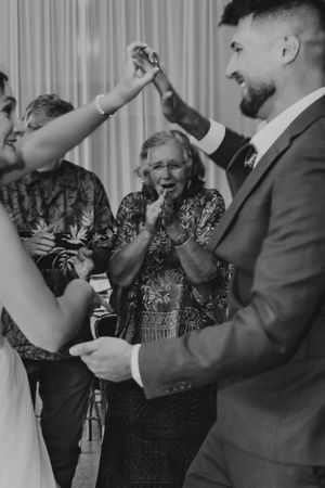 Black and white wedding photo of couple dancing joyfully as elderly guest claps with delight.