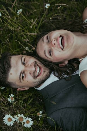 Happy couple laughing while lying in a wildflower meadow with daisies, captured from above in a romantic outdoor portrait.