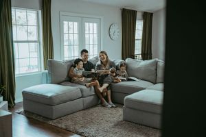 Happy family with children reading a book together on a large gray sectional sofa in a bright living room.
