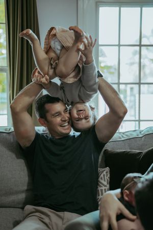Laughing father lifting baby overhead while relaxing on sofa in bright living room.